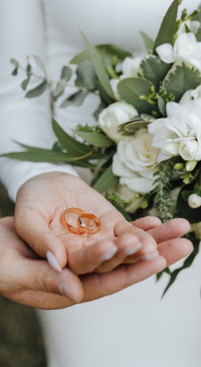 Wedding bands in the hands of bride and groom and with beautiful wedding bouquet made of greenery and white flowers